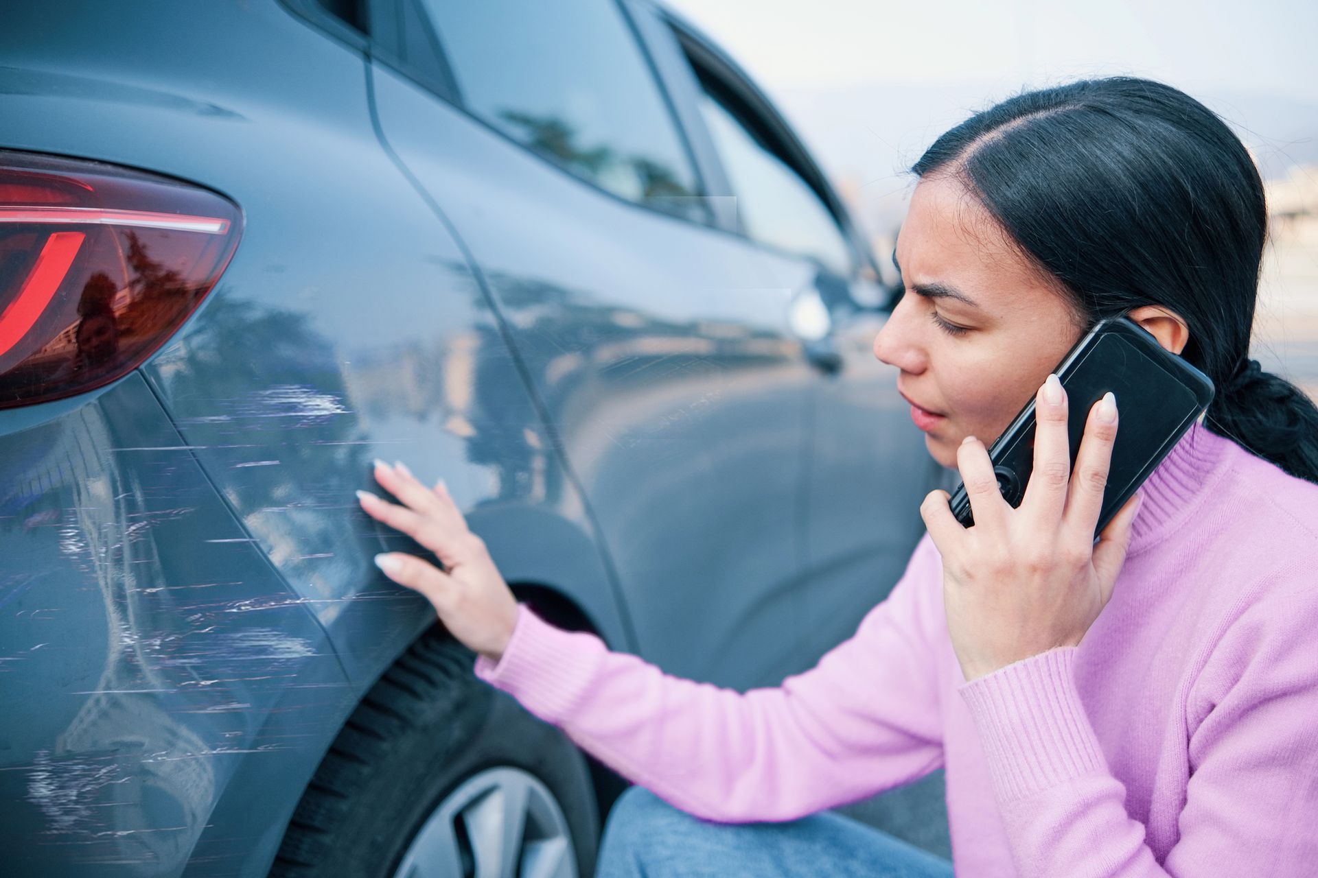 Una persona con un maglione rosa è al telefono mentre tocca un graffio sul lato di un'auto grigia.
