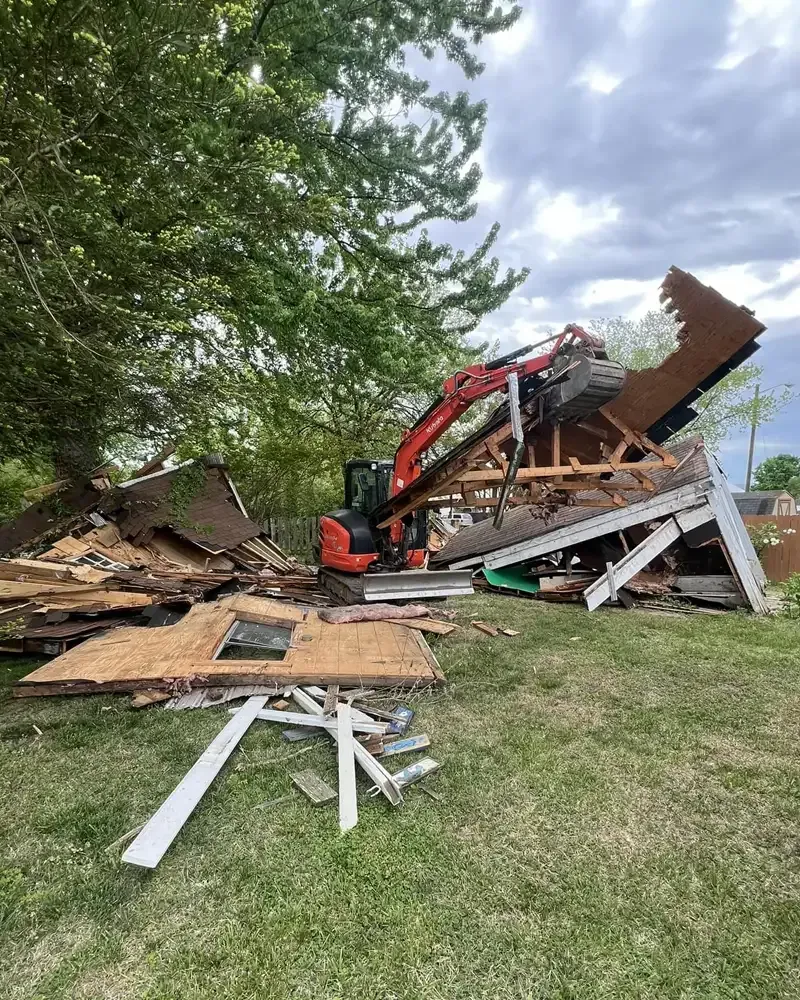 An excavator demolishing a building, scattering debris on green grass under a cloudy sky.