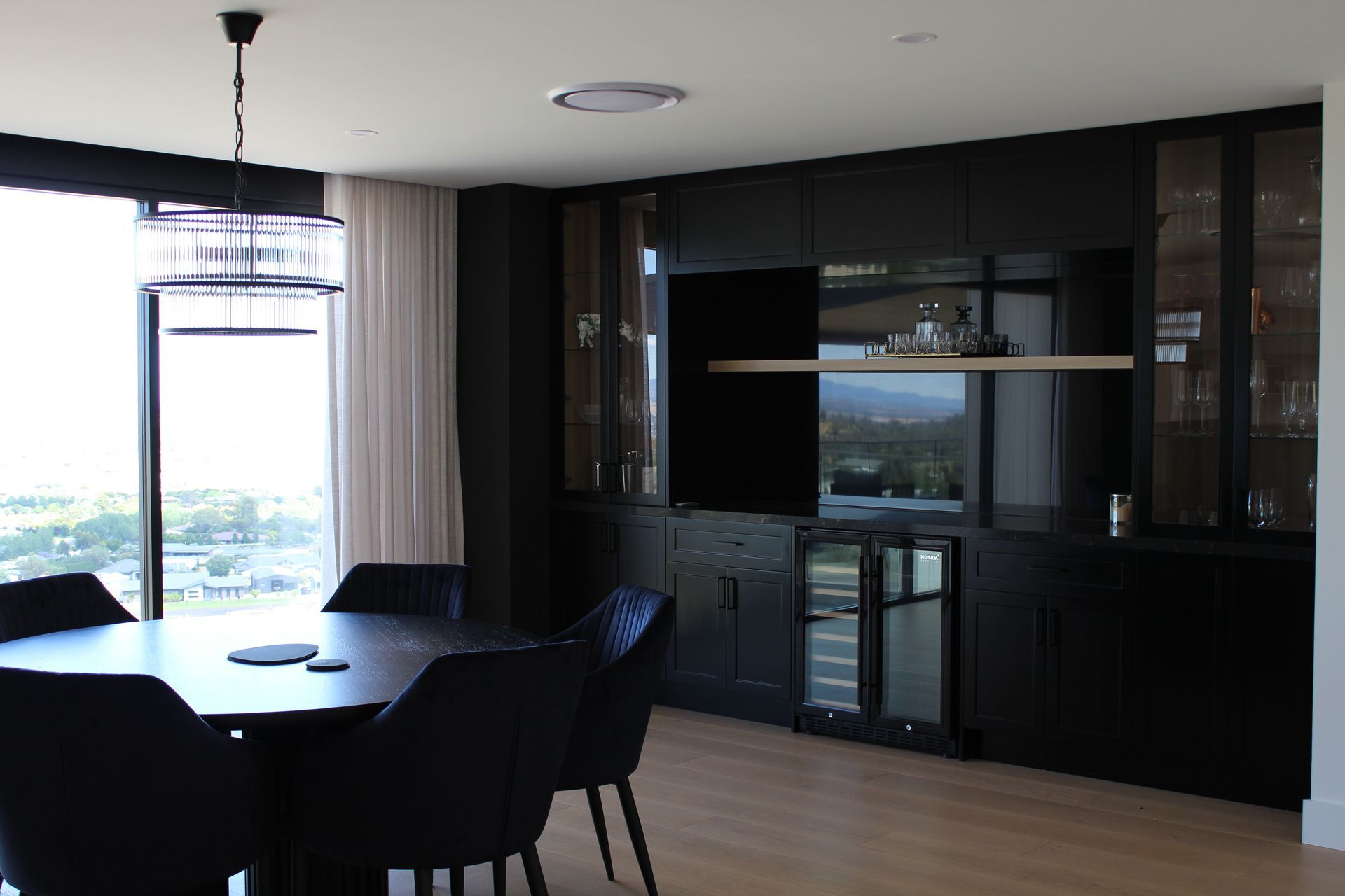 Modern dining area with a round table, dark chairs, and a black kitchen wall with city views.