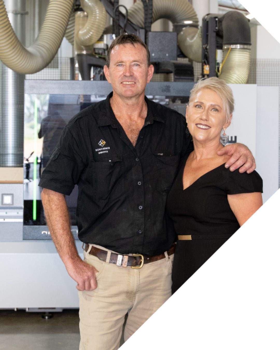 Two people posing in front of industrial equipment, one in a black shirt and beige pants, one in a black dress.