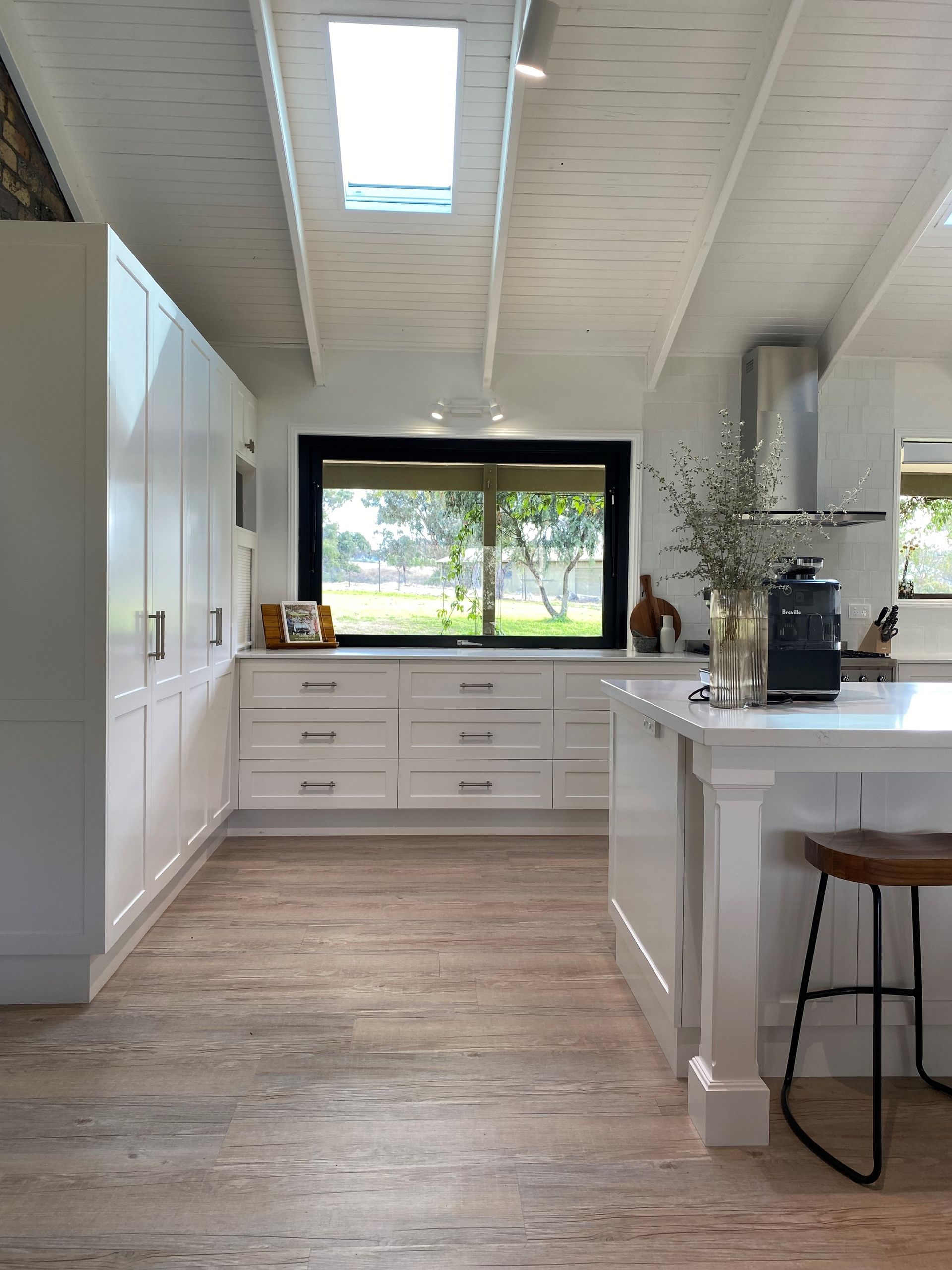 Bright white kitchen with island, tall cabinets, wood floors, and a large window overlooking trees.