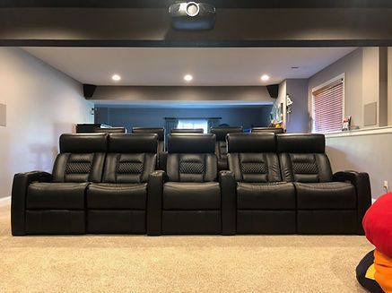 Black leather recliners in a home theater setting. Projector on ceiling, lights, gray walls, beige carpet.