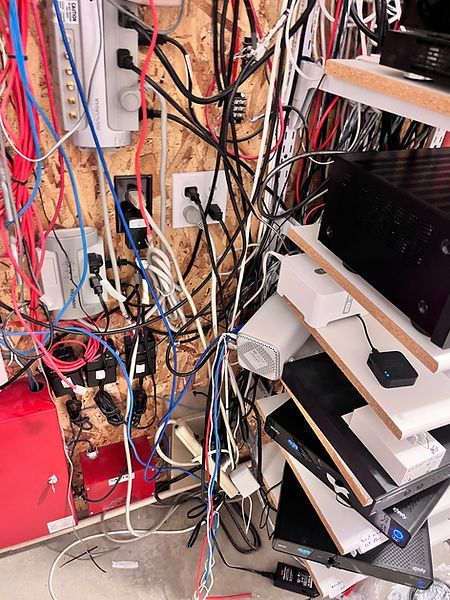 Messy tangle of electrical wires and devices on a wooden wall. Outlets, power strips, and electronics are visible.