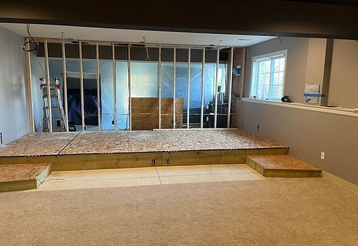 Construction of a stage with steps in a basement, unfinished drywall, exposed studs, and a window.