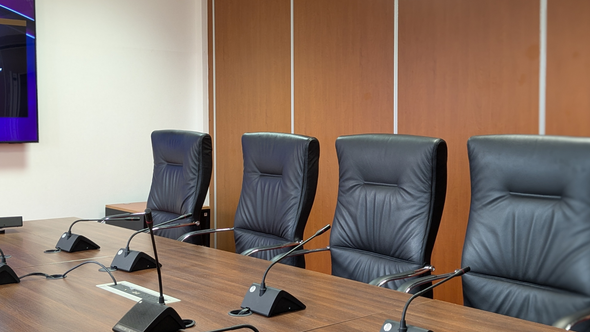 Conference room with leather chairs and microphones, brown wood paneling.