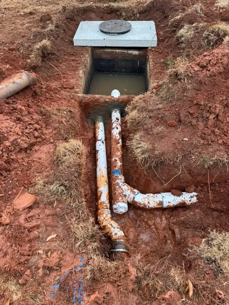 A utility junction box set in red dirt with two white PVC pipes extending from the bottom, one featuring a side branch.