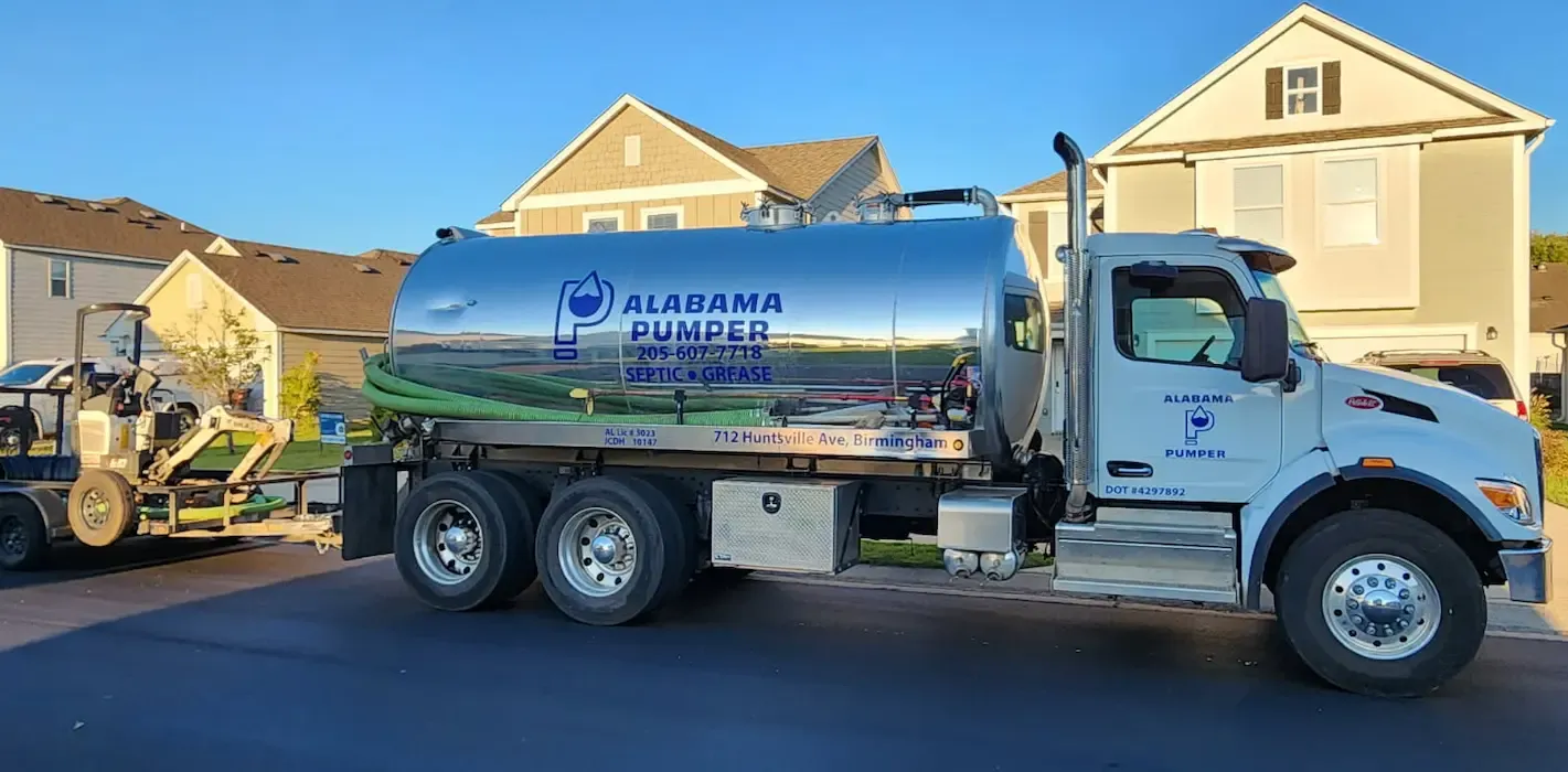 A white Alabama Septic tank truck parked on a residential street in front of houses with a small excavator nearby.
