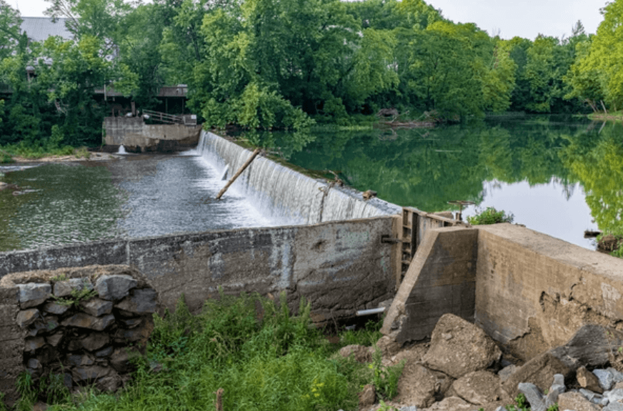 A concrete dam spans a calm river, with water flowing over the center, surrounded by lush green trees.