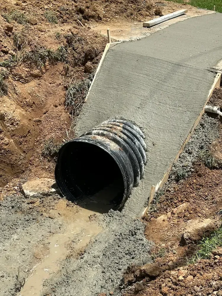 A corrugated black plastic pipe installed into a freshly poured concrete culvert headwall set in an excavated dirt slope.
