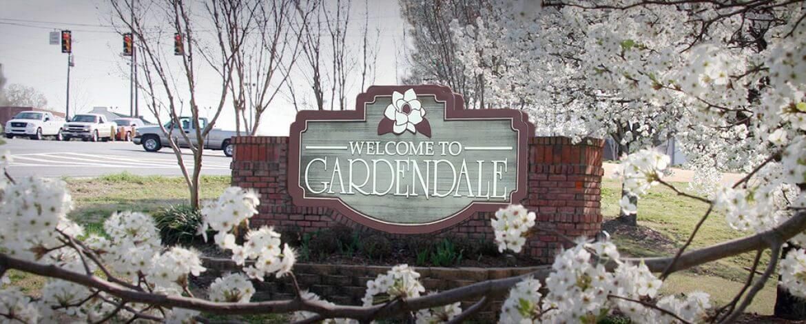 A wooden sign for the city of Gardendale, featuring a flower icon, set in a brick frame surrounded by blooming trees.