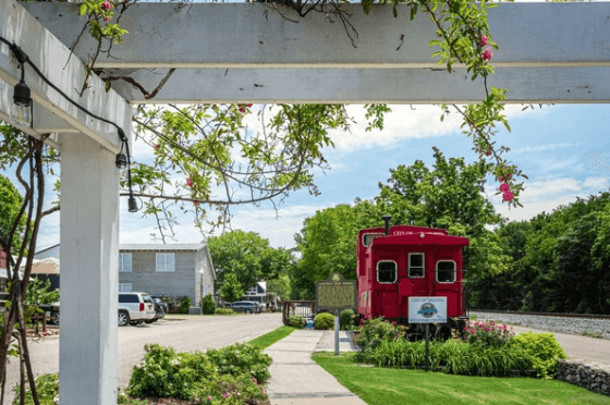 A red train caboose stands on display next to a sidewalk and a white pergola on a sunny, green day.