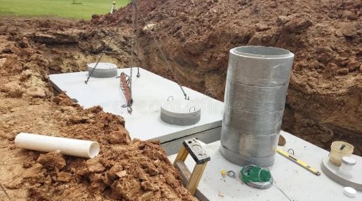 Concrete septic tanks being installed in an excavated dirt trench, with riser segments and tools on-site.