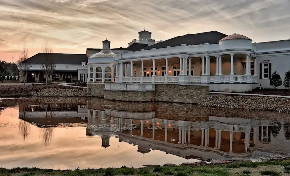 A white, pillared mansion reflected in a calm pond during a sunset with an orange, cloudy sky.