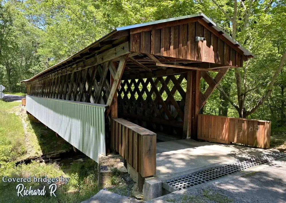 A wooden covered bridge with a lattice truss design stands in a forest setting on a sunny day.