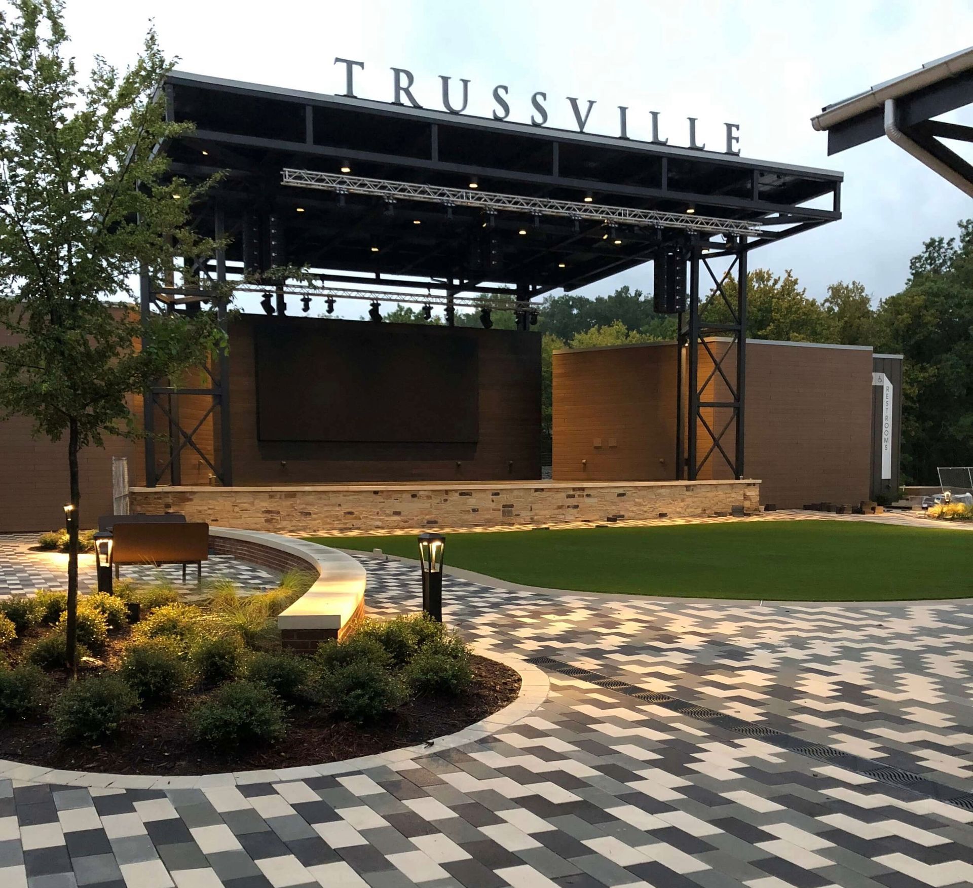 A modern outdoor stage at Trussville with a brick-patterned walkway, green turf, and ambient landscape lighting.