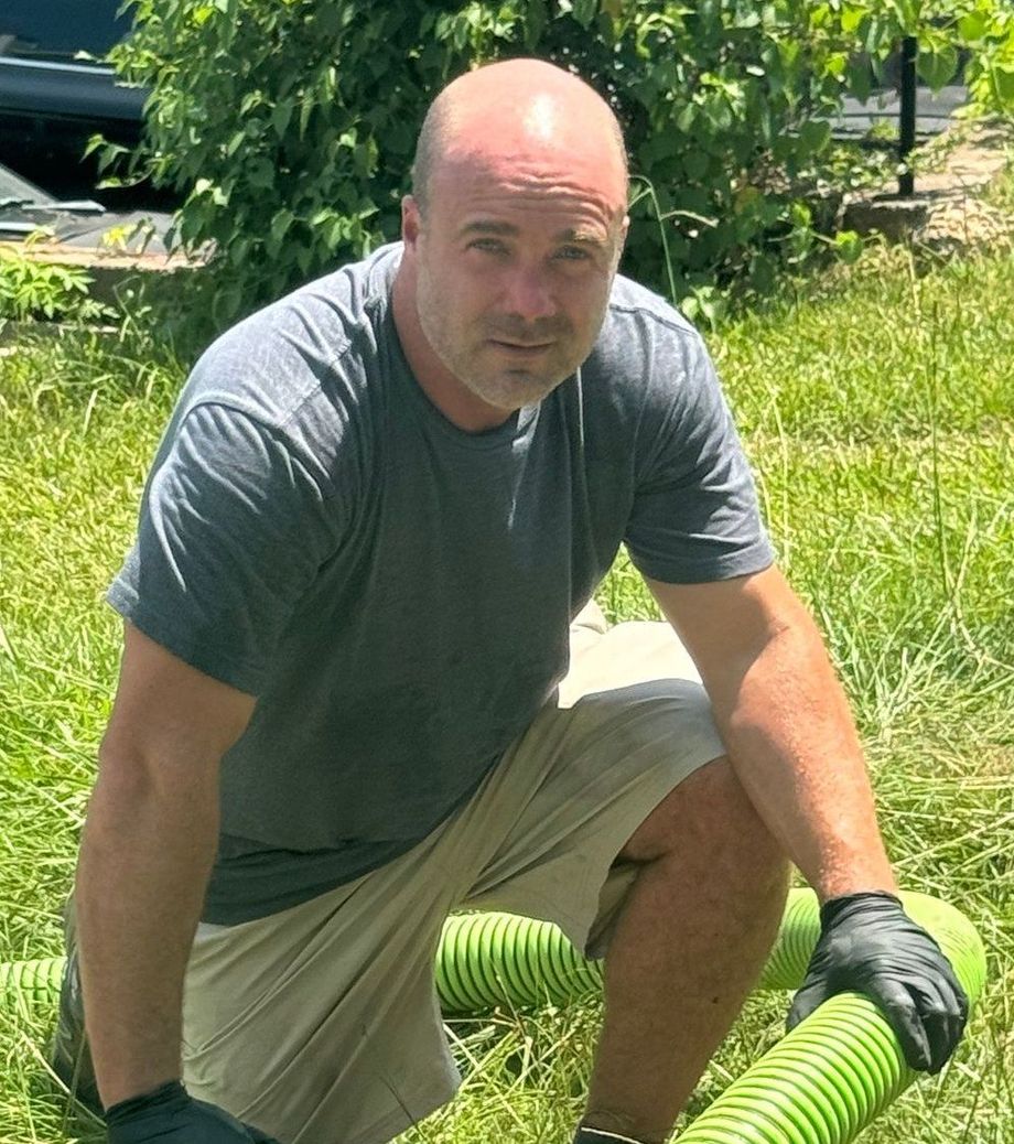 Person kneeling on grass in a gray shirt and khaki shorts beside a bright green hose in a sunny yard