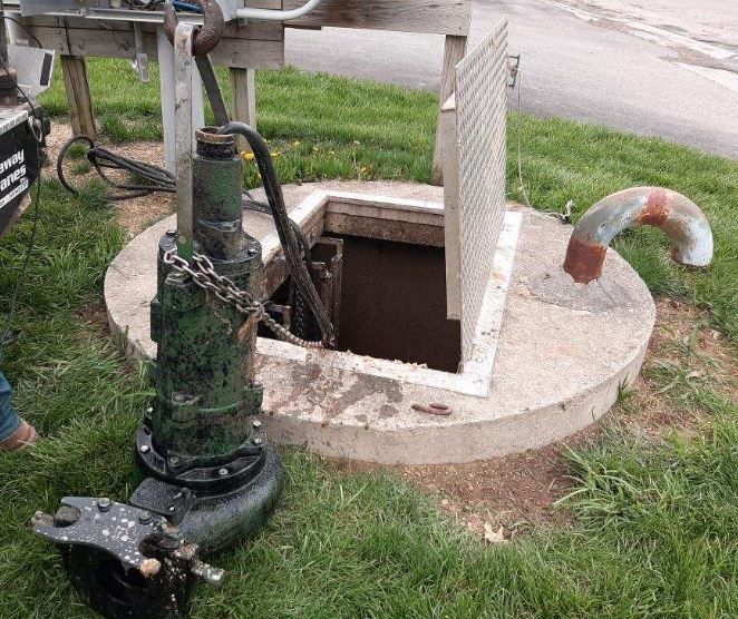 A large, dark green submersible pump sits on the grass beside an open underground maintenance hatch in a grassy area.