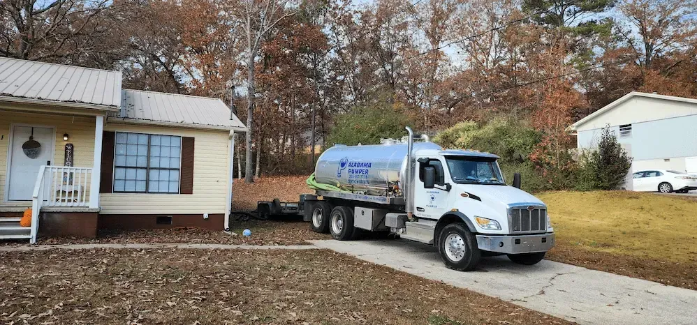 A white septic tank truck parked in the driveway of a beige residential house during autumn.