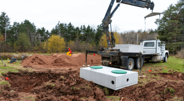 A crane truck lowering a rectangular concrete septic tank into an excavated pit at a construction site.