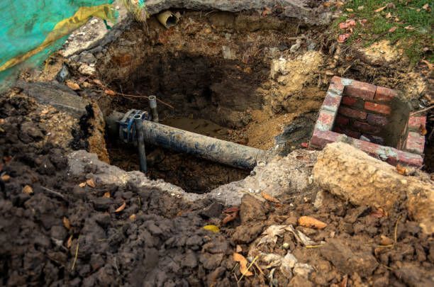 An open excavation pit in dirt reveals a horizontal black pipe with a flange and a nearby small brick structure.