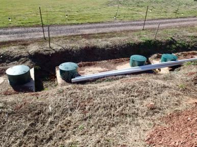 Four green circular septic tank risers installed in a trench on a rural plot, with a long white pipe resting across them.