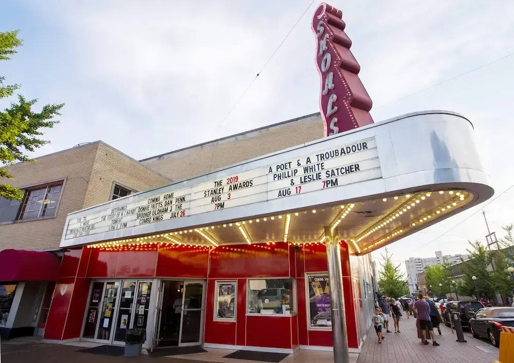The State Theatre with a red marquee and neon signage on a brick building, set on a sidewalk with people walking by.