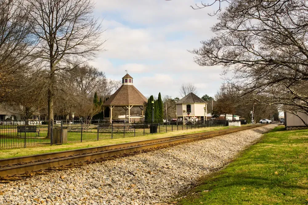 A gazebo sits near railroad tracks and bare trees under a cloudy sky in a park setting.