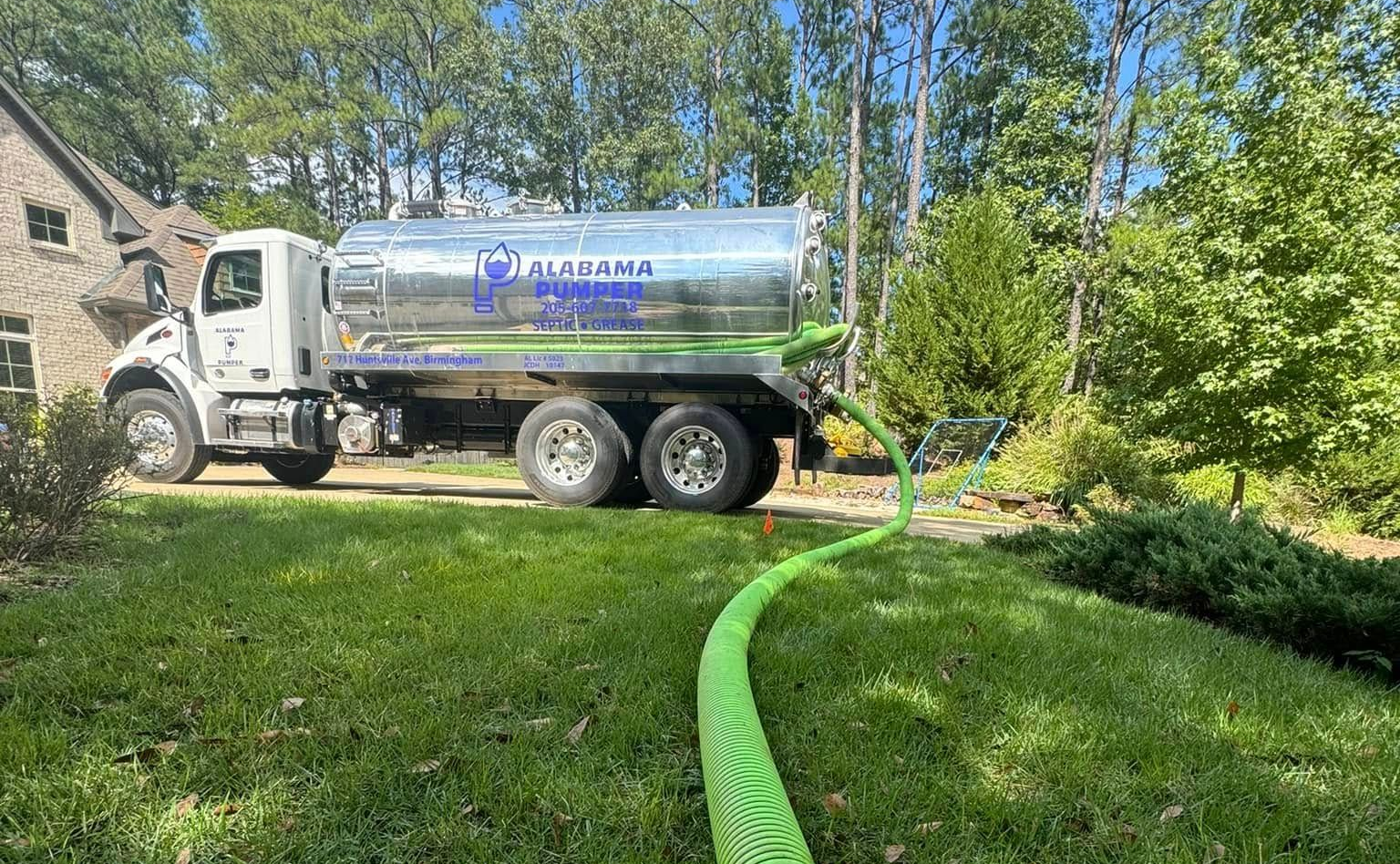 A large truck with a silver tank is parked on a grassy lawn, connected to a long, bright green corrugated hose.