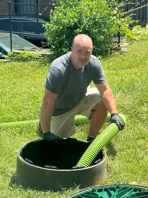 Person kneeling on grass in a gray shirt and khaki shorts beside a bright green hose in a sunny yard