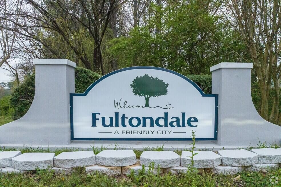 A white, arched welcome sign for Fultondale, Alabama, featuring a green tree logo against a backdrop of trees and shrubs.