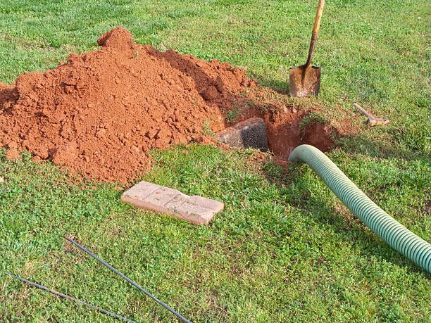 A shovel leans against a large pile of red dirt next to an open hole in a grassy yard, with a green suction hose inside.