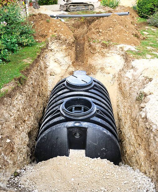 A large black plastic septic tank is installed in a deep dirt trench during construction.