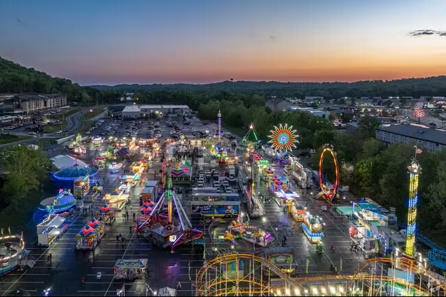 Aerial view of a brightly lit carnival at dusk with various rides and booths set up in a large parking lot.