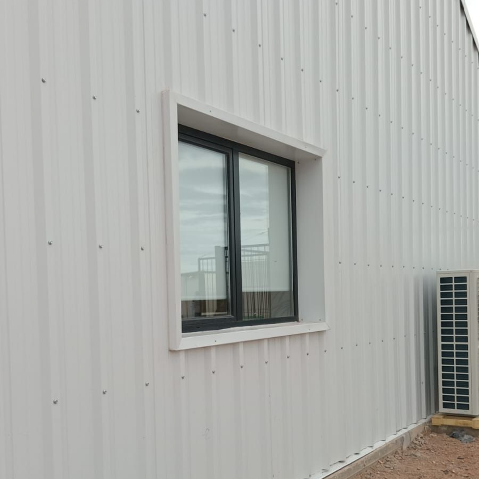 Pared de un edificio de metal corrugado blanco con una pequeña ventana y una unidad de climatización exterior al lado.
