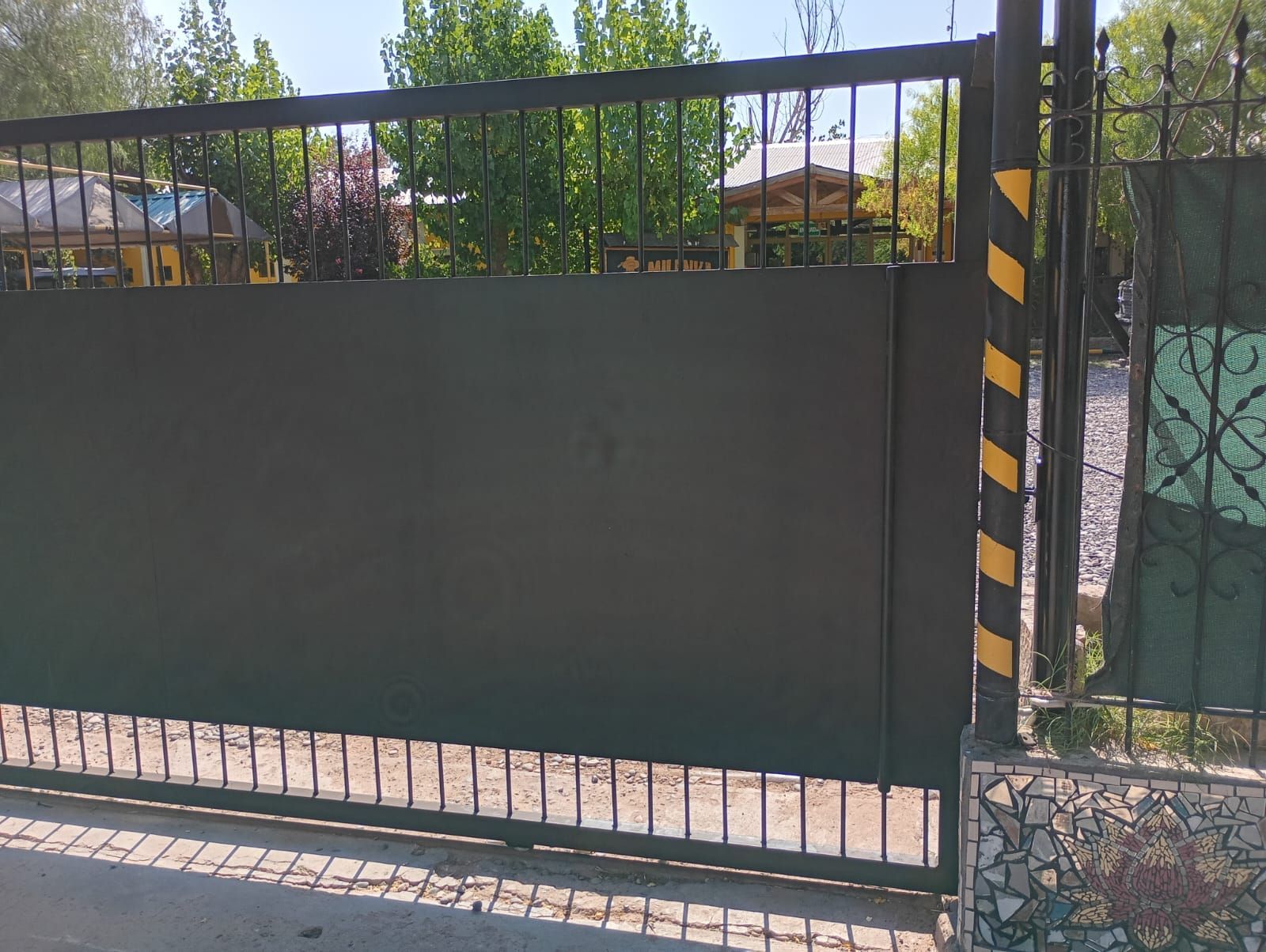 Una verja de metal negro con un gran panel verde oscuro y un poste a rayas amarillas y negras, situada junto a una acera.
