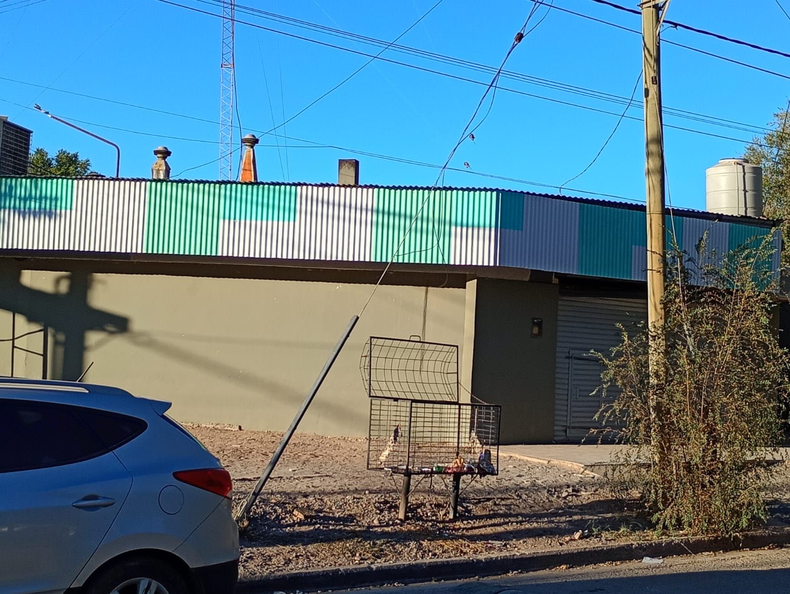 Small beige building with a corrugated roof, parked car, and a wire cart in a sunny lot under a blue sky
