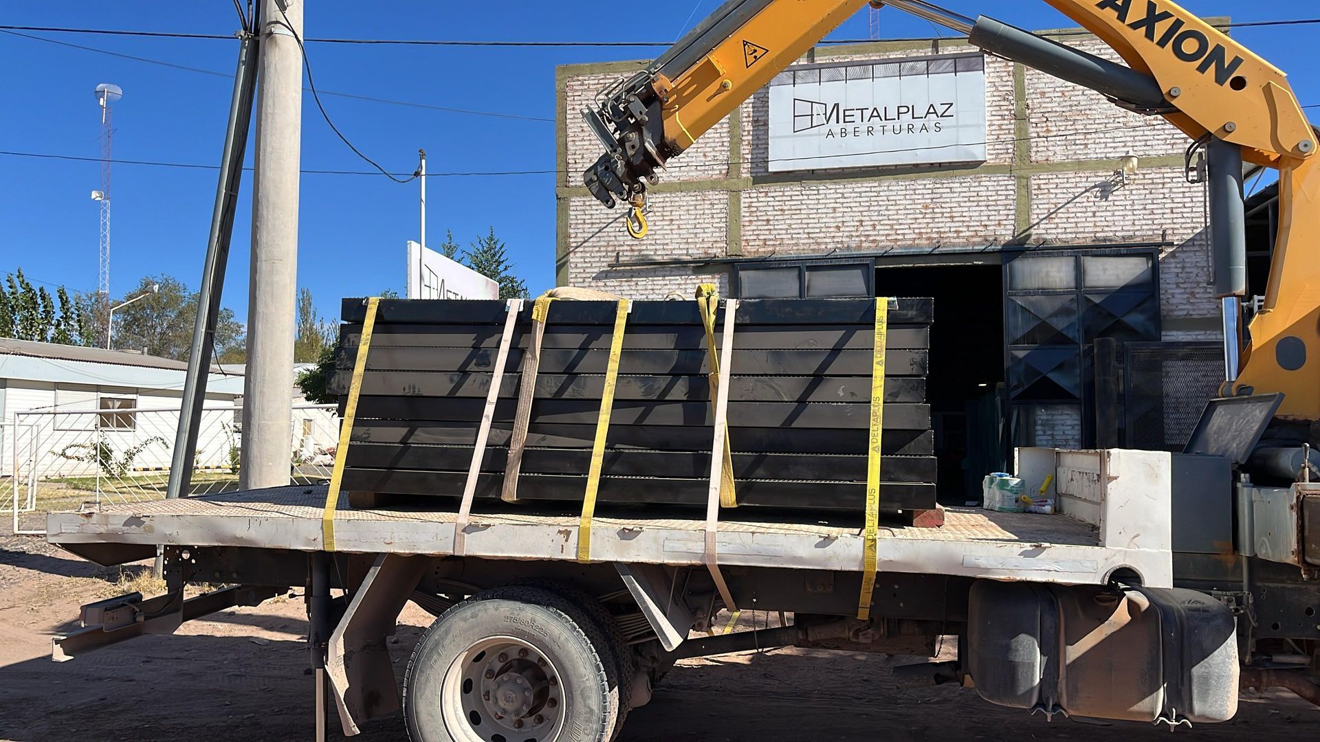 Flatbed trailer loaded with black stacked panels, strapped down with yellow ties, beside an excavator arm outdoors.