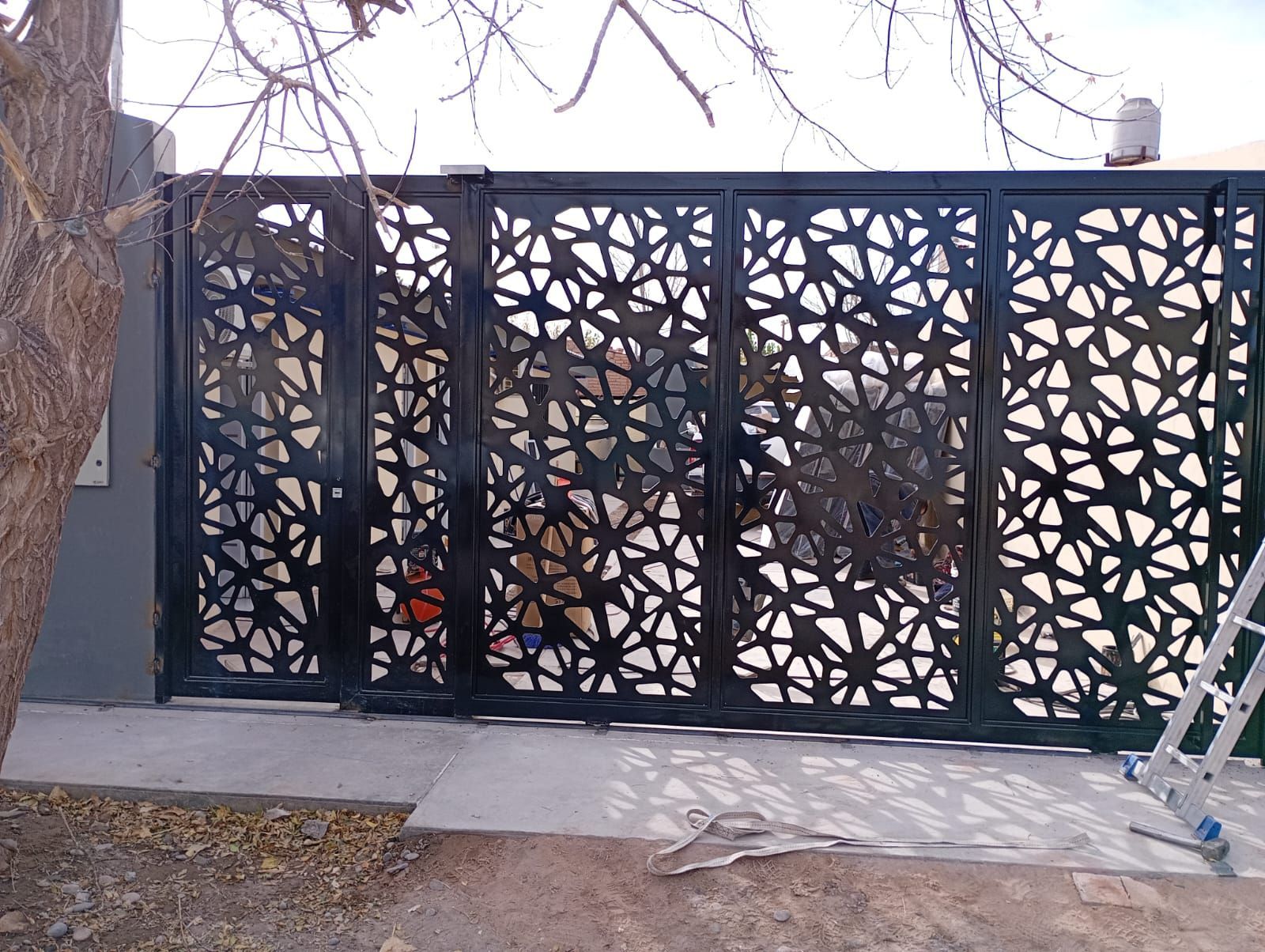 Valla decorativa de celosía negra con estampado geométrico de hojas en la entrada de un patio exterior.