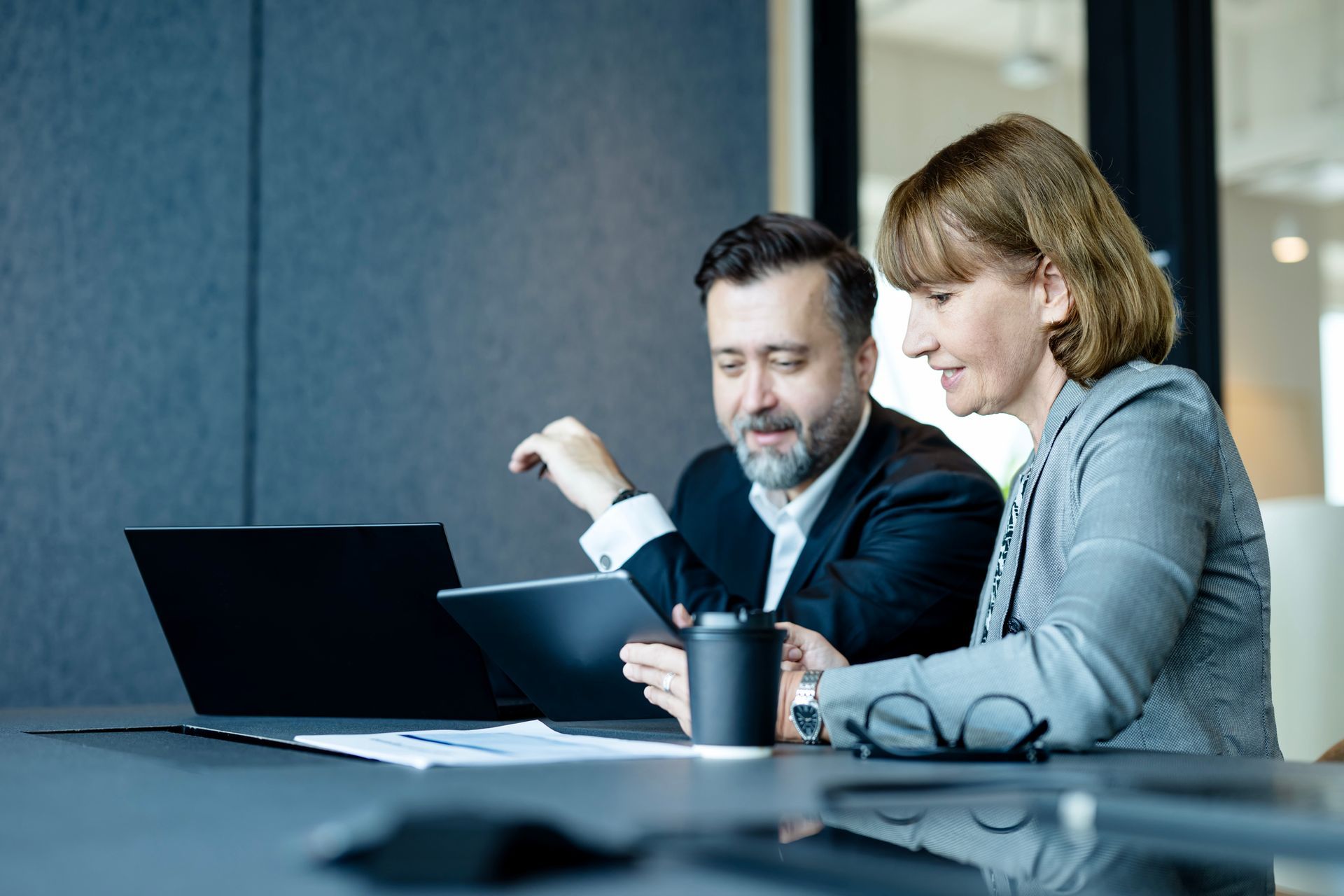 Man and woman in business attire reviewing tablet, black laptop, and coffee at a modern office table.