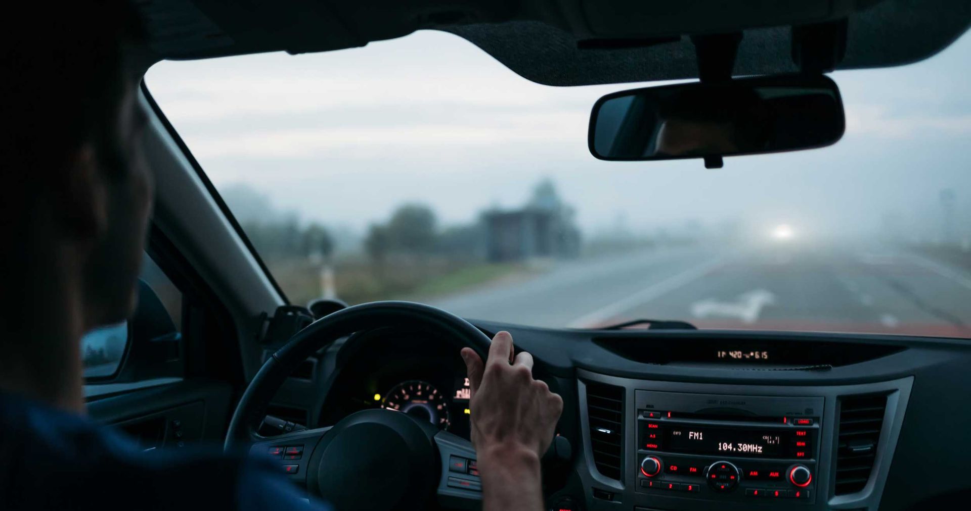 Driver adjusting dashboard controls in a car on a foggy road at dusk