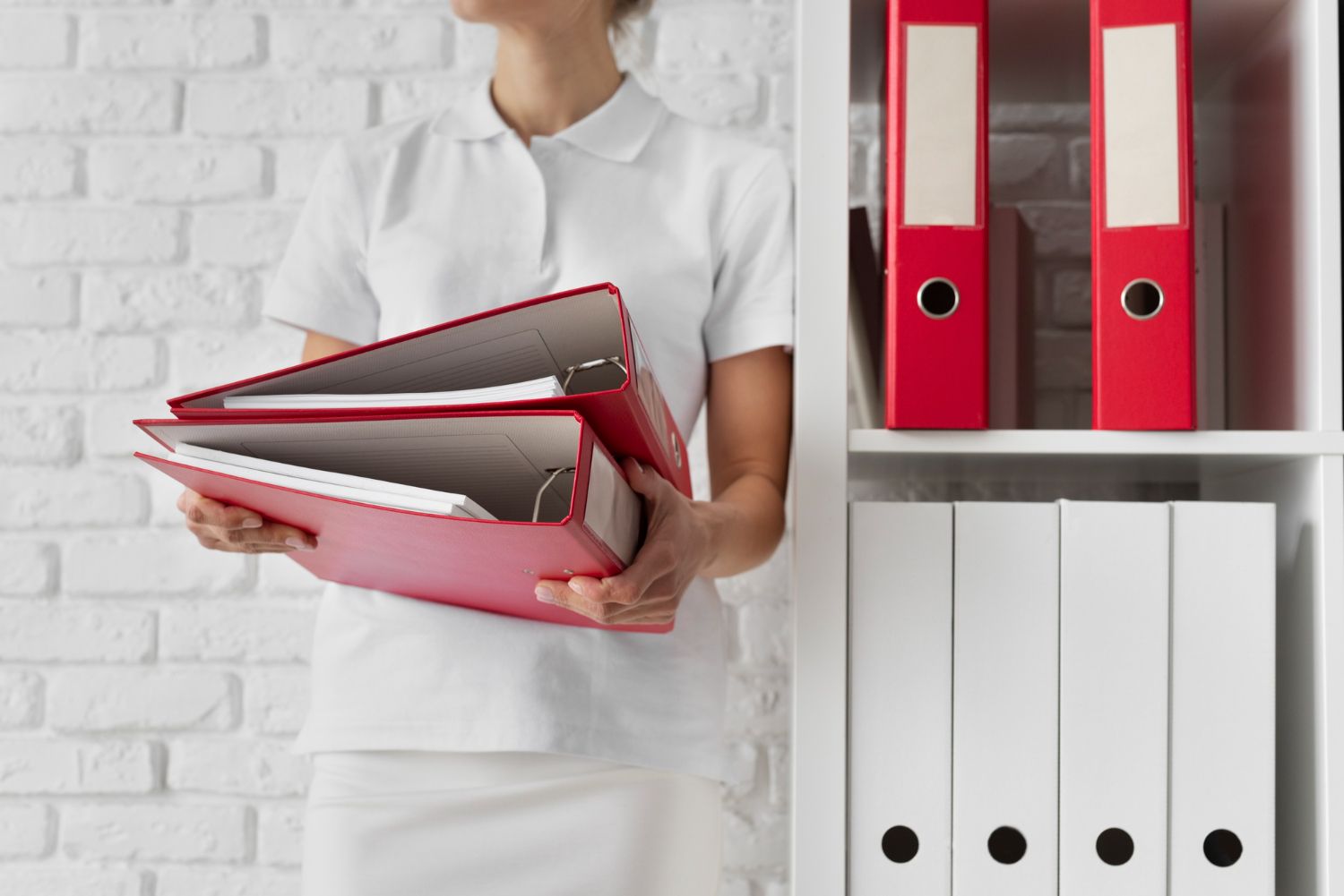 Person holding red and gray binders beside a white shelf with red and white file folders