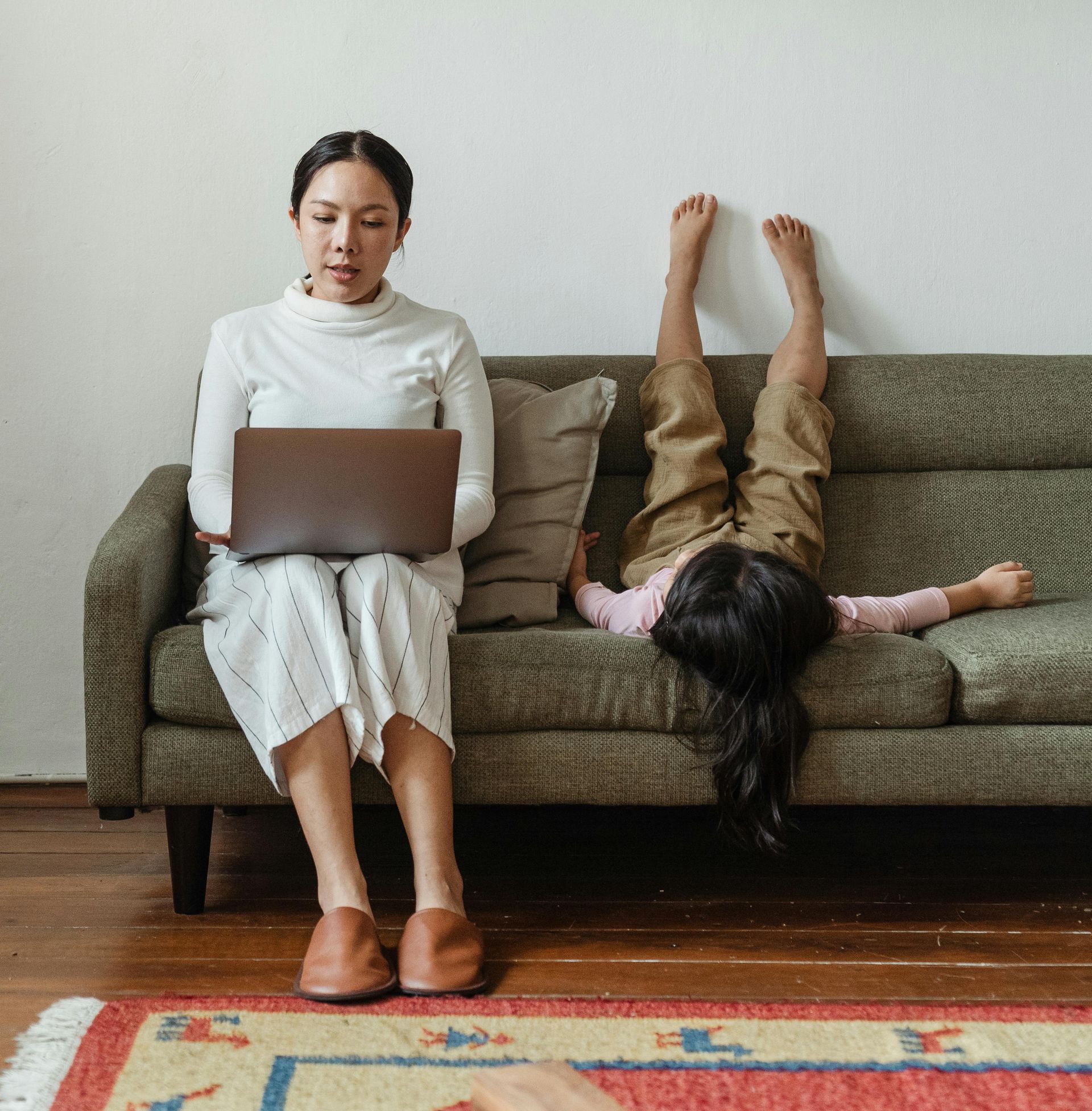 A woman is sitting on a couch using a laptop and a child is laying on the couch.