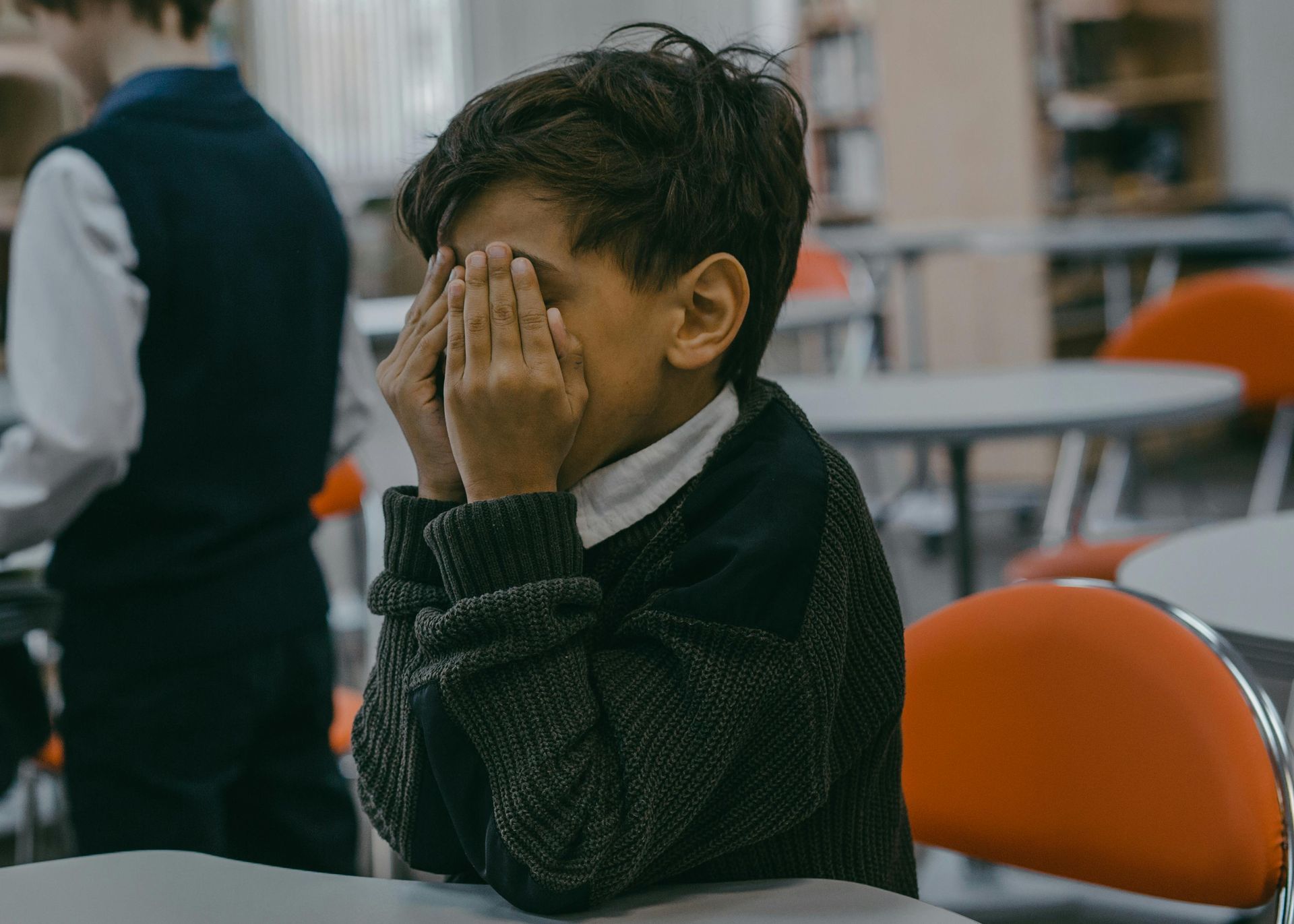 Boy covering his face with his hands, sitting at a table in a classroom.