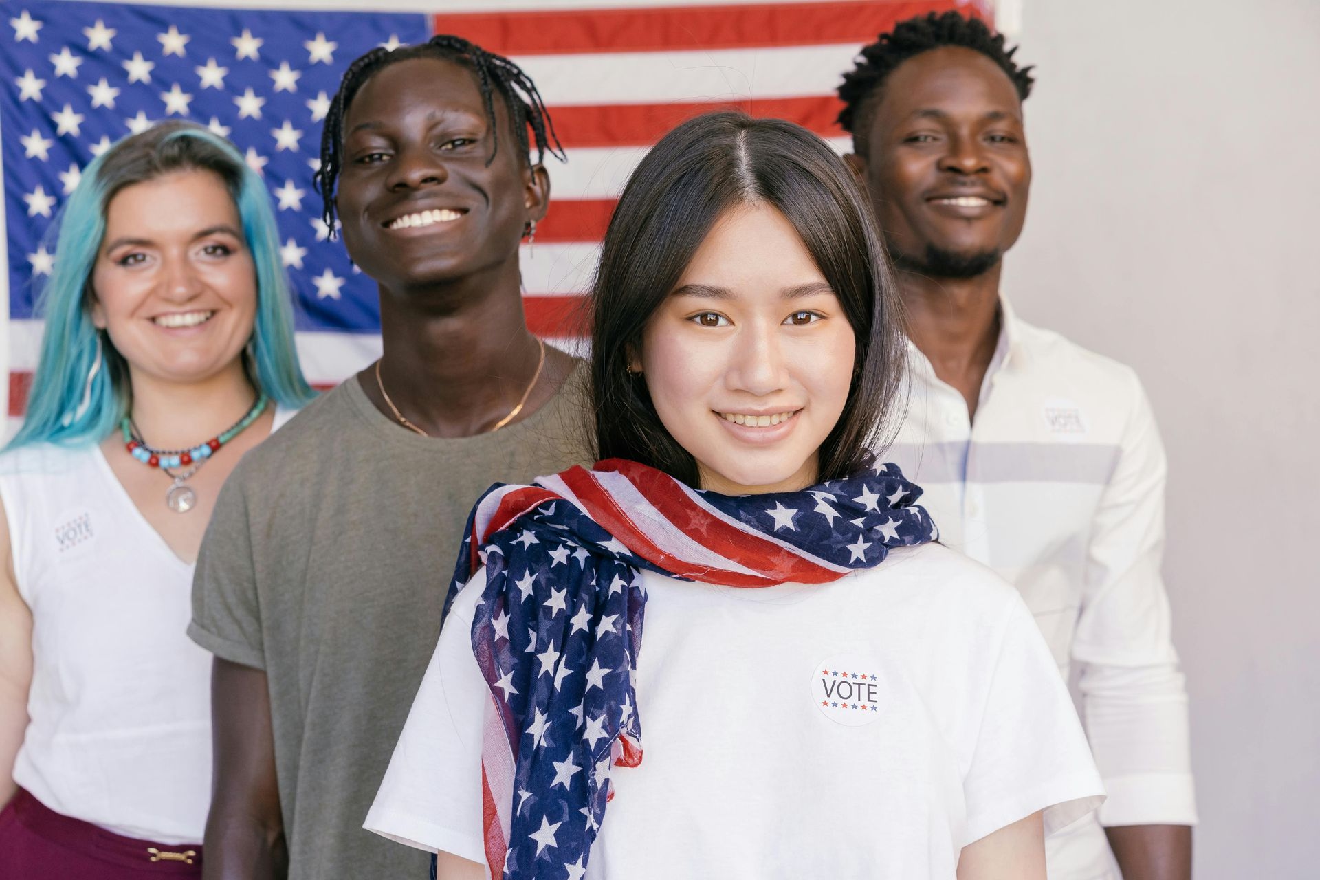 Four people of diverse backgrounds standing in front of an American flag, smiling.