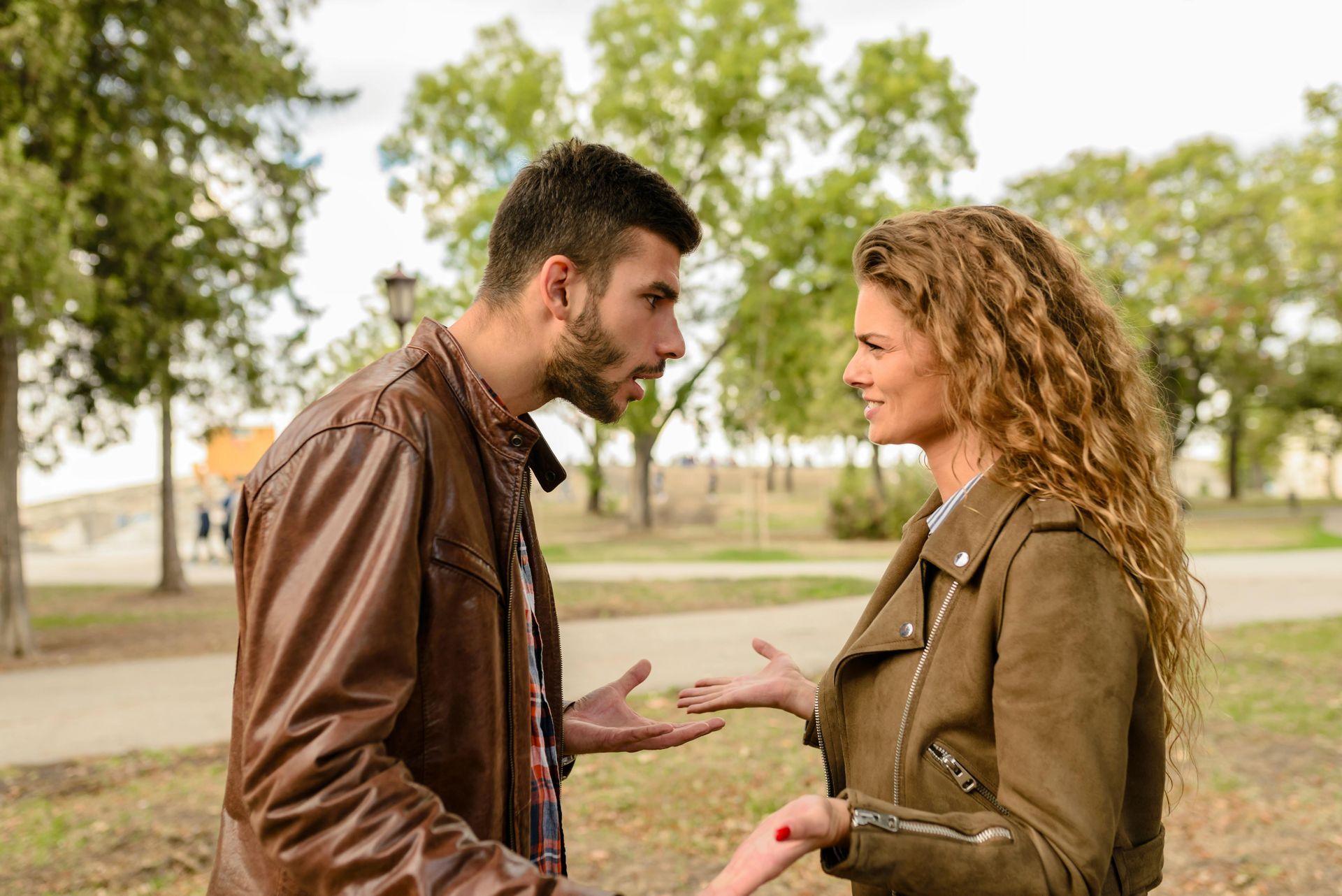 Man yelling at woman in park; both wearing jackets, arguing, man gestures with hands.