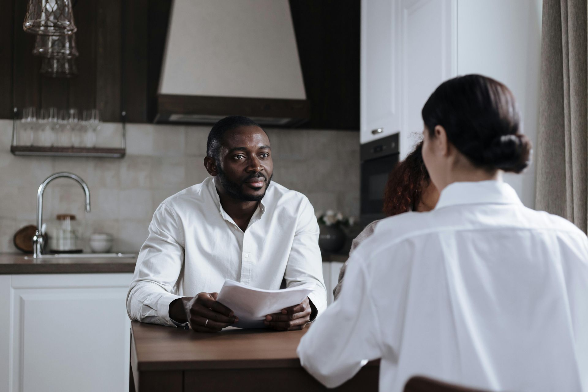 Man holding papers in a kitchen, facing woman. They are both wearing white shirts.