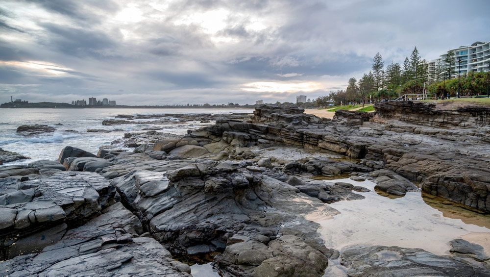 A Rocky Shoreline With A Body Of Water And Trees In The Background — Climatech In Mooloolaba, QLD