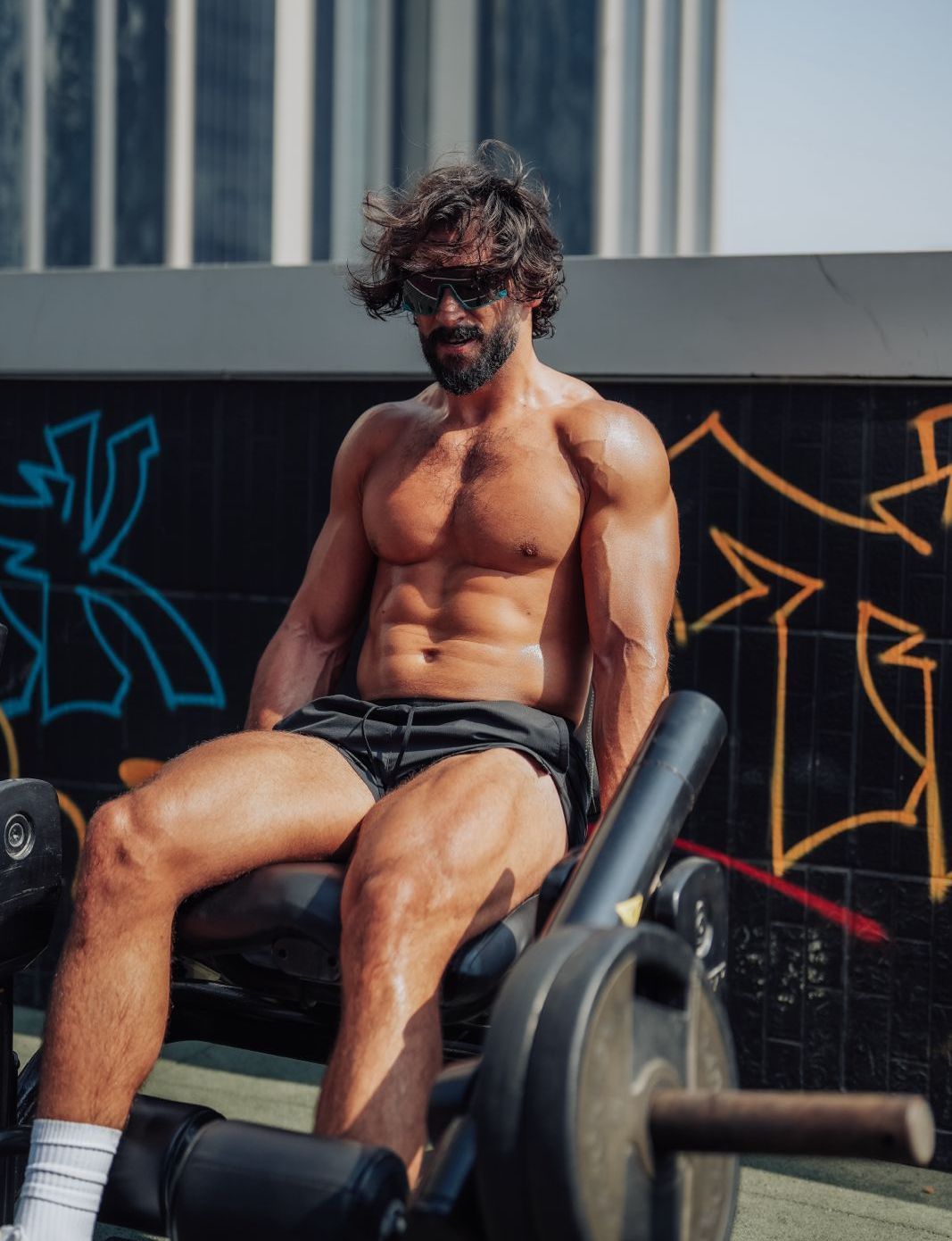 A shirtless man is sitting on a machine in front of a graffiti wall.