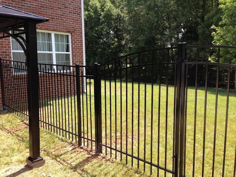 Black metal fence beside a brick house, bordering a grassy yard with trees in the background.