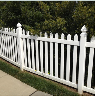 White picket fence along a sidewalk with green trees behind it